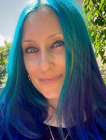 Close-up portrait of a woman with teal blue hair, smiling softly, with greenery and sunlight in the background.