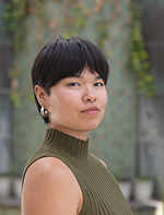 Portrait of a woman with short dark hair wearing an olive green sleeveless turtleneck, outdoors with a blurred garden background.